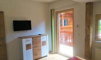 A modern room with a television and a wooden wardrobe. Large windows provide access to a balcony and plenty of daylight.