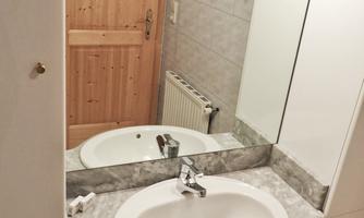 A small bathroom with a round sink and a mirror. The walls are light-tiled and there is a wooden door in the background.