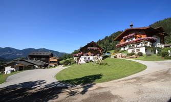 A picturesque mountain landscape with typical Alpine houses and a green meadow. In the background, gentle hills and a clear blue sky can be seen.