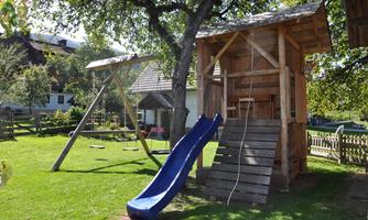 A beautiful playground with a wooden house, a slide, and a swing. The grass is green and sunny.
