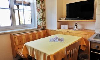 A cozy kitchen corner with a table and bench. A television and windows with curtains create an inviting atmosphere.