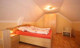 A cozy bedroom with a double bed and light walls. The room has wooden flooring and a modern shelf.