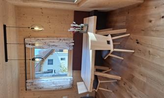 A bright room with wooden paneling and a cozy table with two chairs. A window with curtains offers a view of the neighboring houses outside.