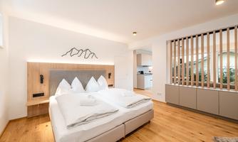 A modern bedroom with a comfortable double bed and wooden floor. In the background, a kitchenette and an elegant wall paneling can be seen.