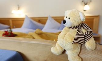 A cozy bedroom with a yellow plush bear on the bed. In the background, cushions and a blanket in warm colors can be seen.