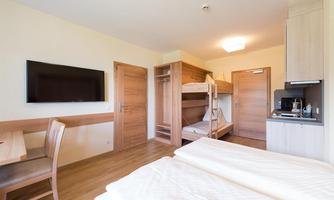 A simple room with a double bed, a bunk bed, and a small kitchenette. The walls are light, and there are wood details as well as a television.