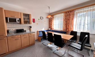 A modern kitchen with wooden cabinets and a large dining table. Large windows provide plenty of light and a welcoming atmosphere.