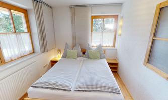 A cozy bedroom with a double bed and bright windows. The walls are painted in neutral colors and the floor is made of wood.