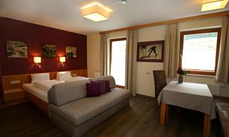 A cozy hotel room with two beds, a sofa, and a table. The walls are painted in warm colors, and two windows provide daylight.