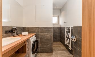 A modern bathroom with a washing machine and a wooden washstand. The walls are tiled in gray, and there is a window for natural light.