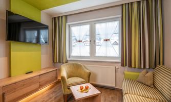 A modern room with a TV on the wall and a cozy seating area. The windows are decorated with colorful curtains and a table with fruit stands in the middle.