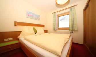 A cozy bedroom with a double bed and light pillows. The room has a window with curtains and simple wooden furniture.