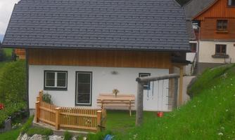 A modern house with a wooden veranda and a swing. Surrounded by green grass and mountains.