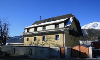 A yellow house in the snow with a dark roof and shutters. In the background, there are mountains and a clear blue sky.