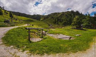 A green meadow with a gravel path and hills in the background. The sky is cloudy, but the landscape looks inviting and calm.