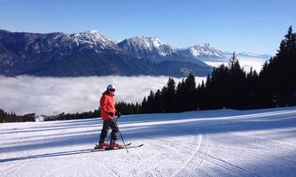 A skier is riding a slope with snow-covered terrain. In the background, impressive mountains and a clear blue sky can be seen.