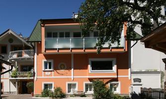 A beautiful residential building in orange with large windows and a modern balcony. Well-maintained plants surround the house and a blue sky is visible in the background.