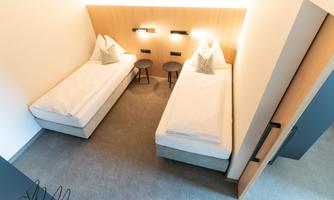 A modern room with two single beds and a minimalist design. The walls are light and illuminated with warm light.