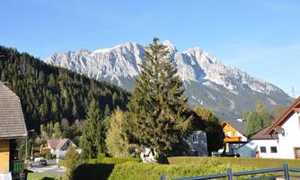 A picturesque mountain landscape with tall mountains and green forests. In the foreground, there are some houses and trees.
