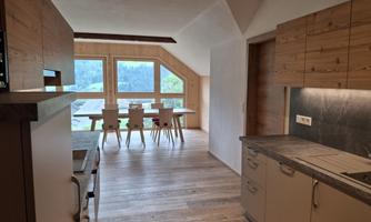 A modern kitchen with wooden cabinets and an open living area. Large windows offer a view of the surrounding landscape.
