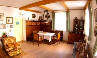 A cozy room with a wooden ceiling, a large table, and various chairs. Pictures hang on the walls, and a comfortable couch is in the corner.