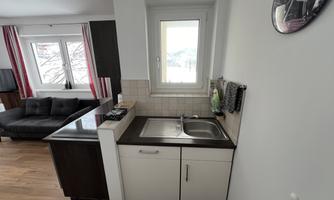 A modern kitchen with a simple, functional design. The window offers a view outside and the living area is well visible.