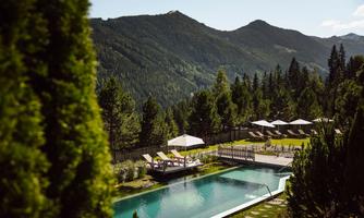 A beautiful pool in the midst of a green garden, surrounded by mountains. Relaxation areas with loungers and parasols create an inviting atmosphere.