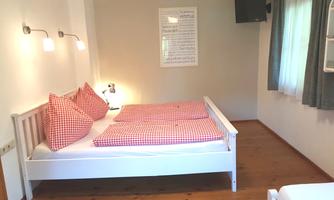 A cozy bedroom with two single beds and red pillows. There is a television on the wall and a bright atmosphere from the window.