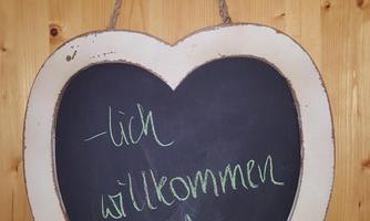 A heart-shaped signboard with the inscription "Welcome" and a small tree drawing. The sign is hanging on a wooden wall.