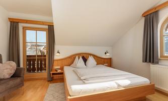 A cozy bedroom with a large bed and carefully arranged pillows. The room has a window overlooking a balcony and is bright and inviting.