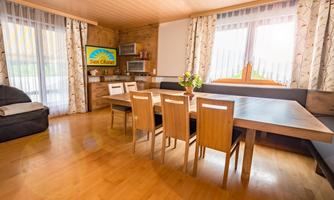 A bright living room with a large table and six chairs. The windows let in plenty of light, and there is a television with a sunny motif.