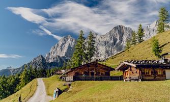 A picturesque mountain landscape with high peaks and a clear sky. In the foreground, there are traditional wooden chalets and a narrow hiking trail.