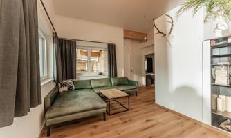 A modern living room with a green sofa and a wooden table. Large windows provide plenty of daylight and a cozy atmosphere.