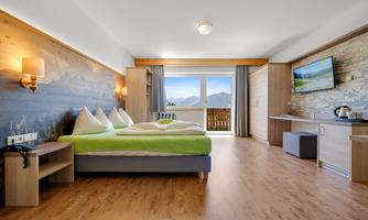 A modern hotel room with a large window that looks out at the mountains. The bed has green bedding and the walls are decorated with wood paneling.
