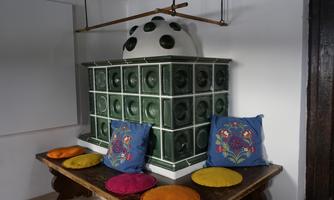 A traditional tiled stove in green stands in a room. Colorful cushions lie on a wooden bench in front of it.