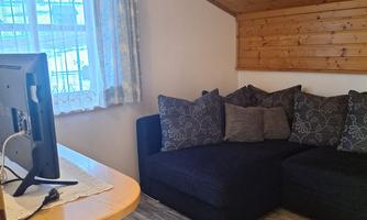 A cozy living space with a black sofa and several cushions. Next to the sofa, there is a television on a table, and the light comes through a window with curtains.