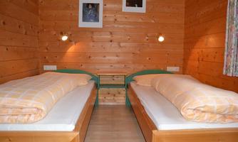 A cozy room with two single beds and wooden paneling. Two pictures hang on the wall, and the light is warm and inviting.
