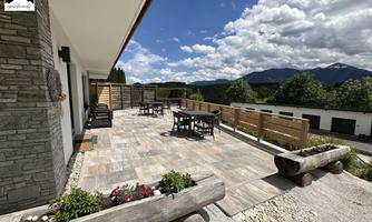 A spacious terrace with wooden furniture and a view of the mountains. The sky is clear and full of clouds.