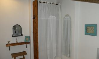 A bright room with a wooden chair and a small wall shelf. A curtain separates the area, while a mirror and a decorative frame hang on the wall.