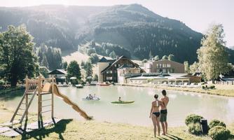 A beautiful lake surrounded by mountains, with a slide area in the foreground. Two people stand on the shore and watch the water.