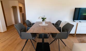 A modern dining area with a wooden table and comfortable chairs. There is a plant on the table and in the background, a television is visible.
