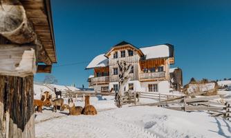 A cozy farmhouse in a snowy landscape. In the foreground, several animals stand in the snow, enjoying the clear weather.