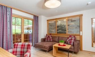 A cozy living room with a modern couch and a wooden table. Large windows let in plenty of light and provide a view of the outdoors.