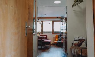 A modernly furnished hallway with a door leading to a bright living area. In the background, tables and chairs are visible.