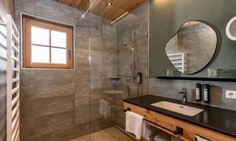 A modern bathroom with a glass shower and a rectangular mirror. The room is designed with gray tiles and wood details, and the wash area contains skincare products.
