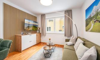 A modern living room with a cozy couch and a TV on the wall. The room has large windows and warm wood decor.