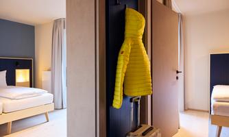A bright room with two beds and an open door. A yellow jacket hangs on the wall, while a suitcase stands on the floor.