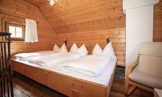A cozy wooden bedroom with a double bed and white bedding. A chair and windows create a bright, inviting atmosphere.