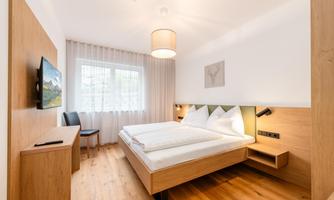 A modern bedroom with a double bed and a TV. Large windows provide ample daylight and a pleasant atmosphere.