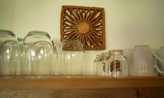 A shelf with glasses and decorative vessels. In the background, there is an artistic wall ornament.
